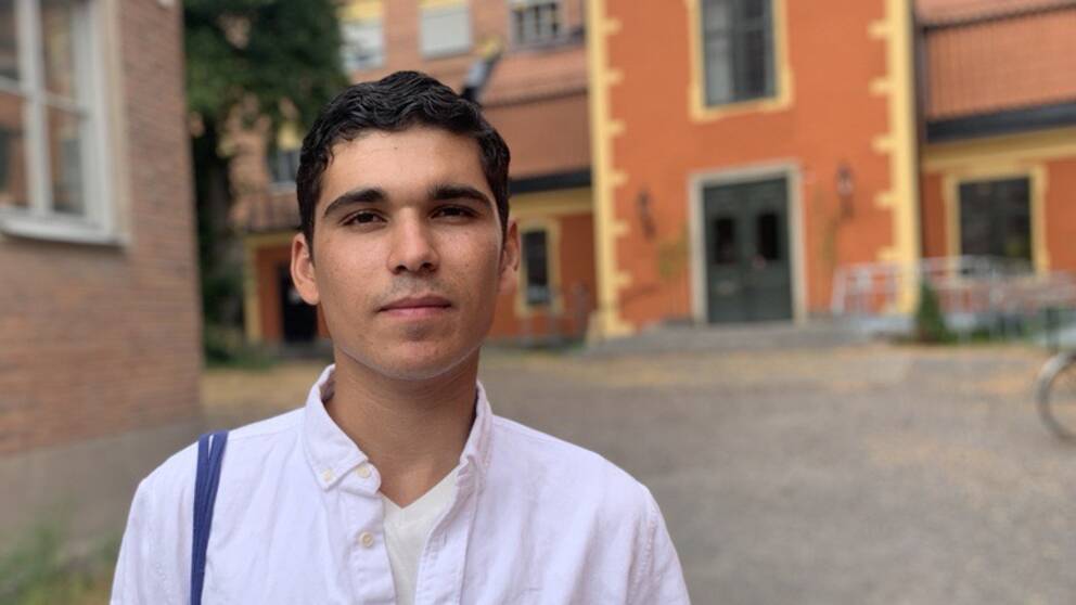 Man, vit skjorta, mörkt hår, i bakgrunden oranget hus.