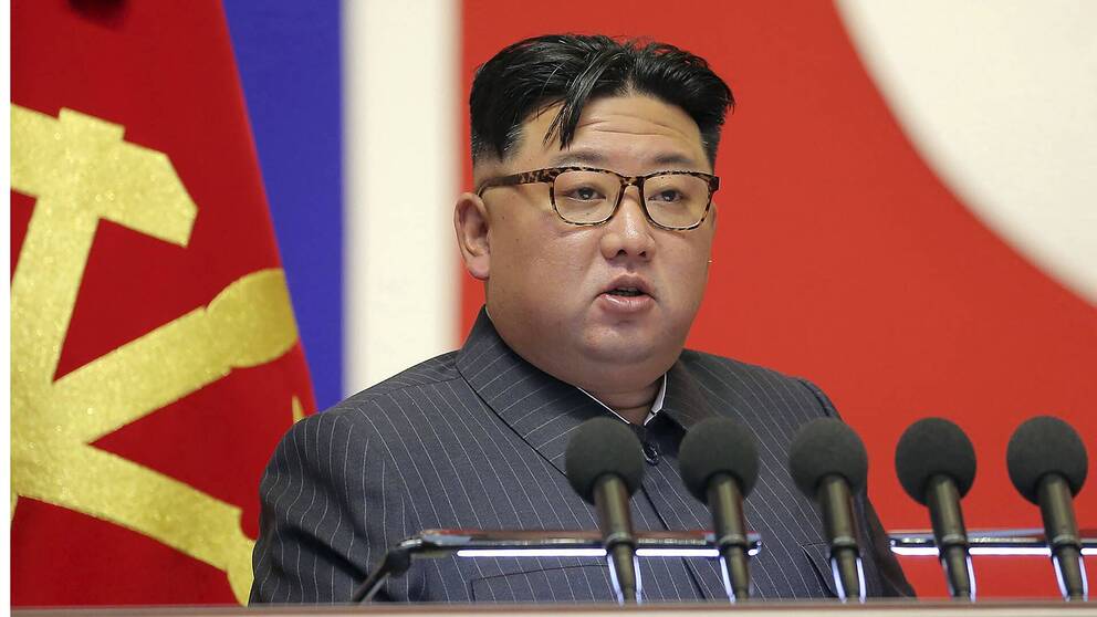 Nordkoreas ledare Kim Jong Un sitter framför en nordkoreansk flagga och flera mikrofoner och håller tal.