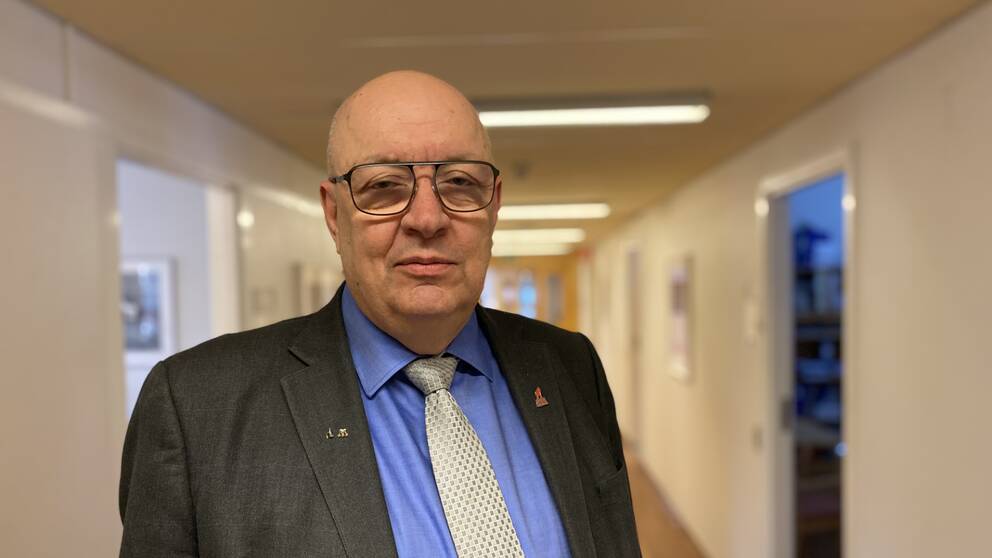 En man (politiker vid namn Ulf berg (M)) har glasögon och kostym och tittar in i kameran – bakom en korridor
