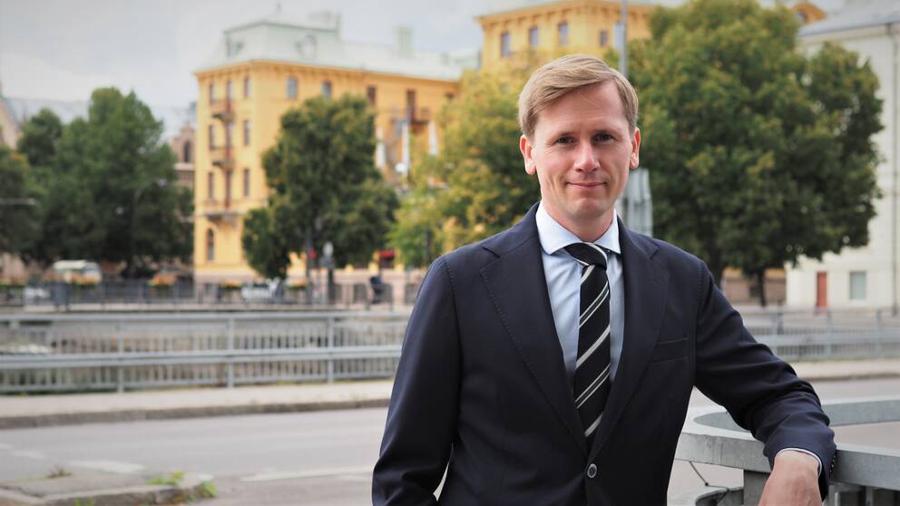 Roger Hedlund – en man i kostym och slips vid räcke, gata och stor äldre tegelbyggnad i bakgrunden