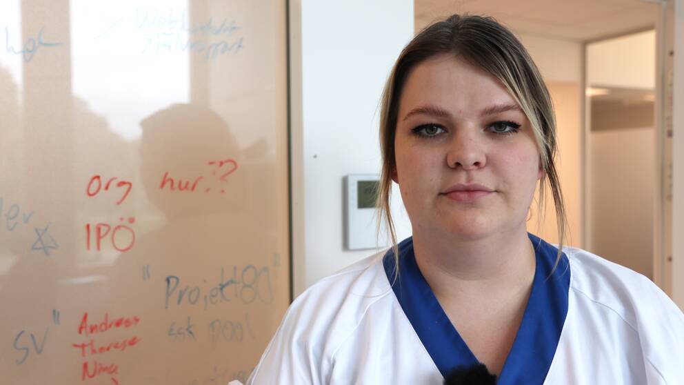Halvbil på Elsa Jäger, ler något, mörkblont hår, grönbruna ögon, ljus hud, vit arbetsskjorta. I bakgrunden, vänster ett glasfönster och till höger en dörröppning.