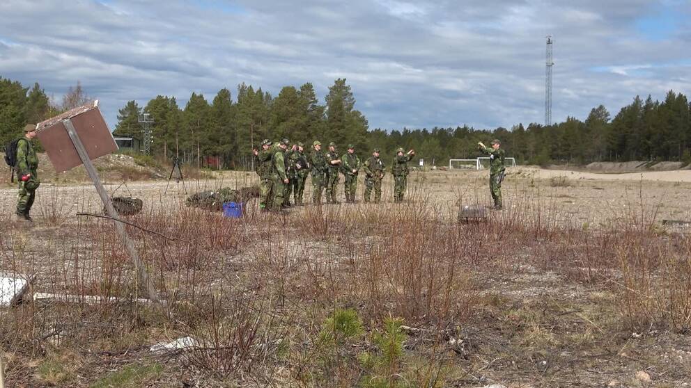 en grupp människor i gröna kamouflagekläder står på en öppen yta, skog i bakgrunden.