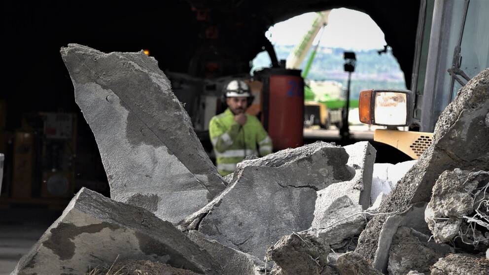 biter av riven betongstruktur, i bakgrunden en person i arbetskläder och hjälm framför en tunnelöppning