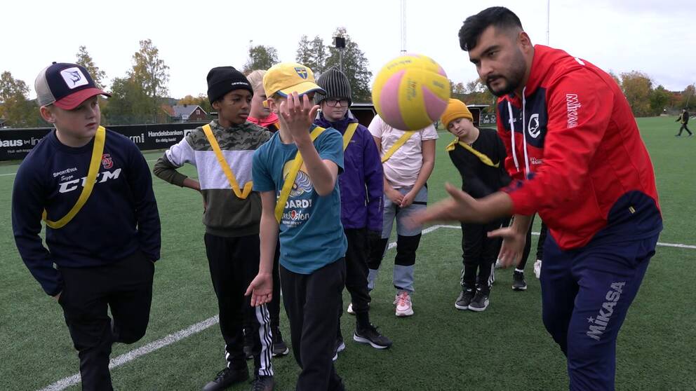 En tränare med röd jacka visar några barn hur man servar med en Volleyboll, bollen är i luften.