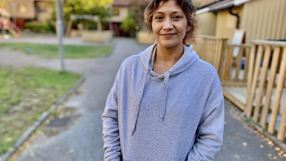 En kvinna med mörkt hår iklädd grå hoodie står i ett bostadsområde.