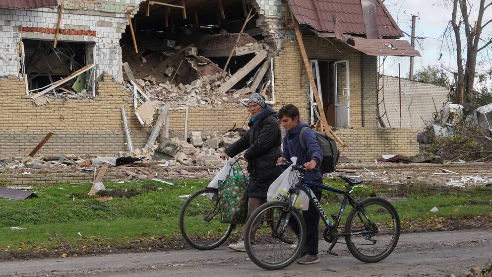 En vuxen och ett barn styr sina cyklar föbi ett förstört hus i den ukrainska byn Petropavlivka.