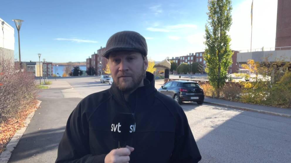 Reporter Emil Hammarström står framför kameran och berättar om händelsen, bakom honom står en mörk parkerad bil och man ser en ett grönt träd till höger om Emil som bär en basker och mörk jacka.