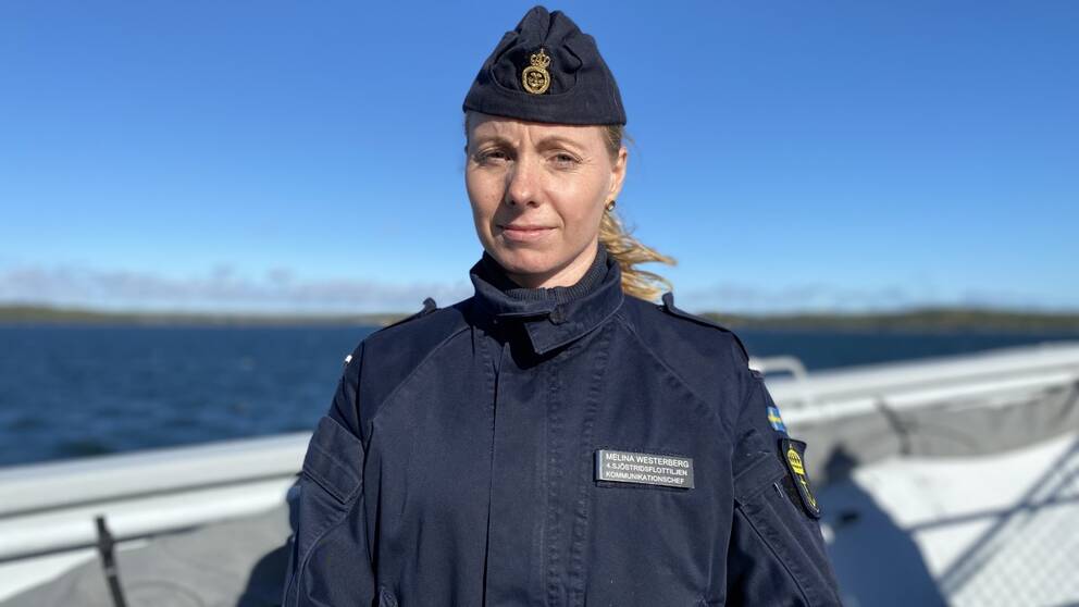 Melina Westerberg från försvarsmakten står på en båt i skärgården med blå bakgrund. Hon har uniform och kisar mot solen.