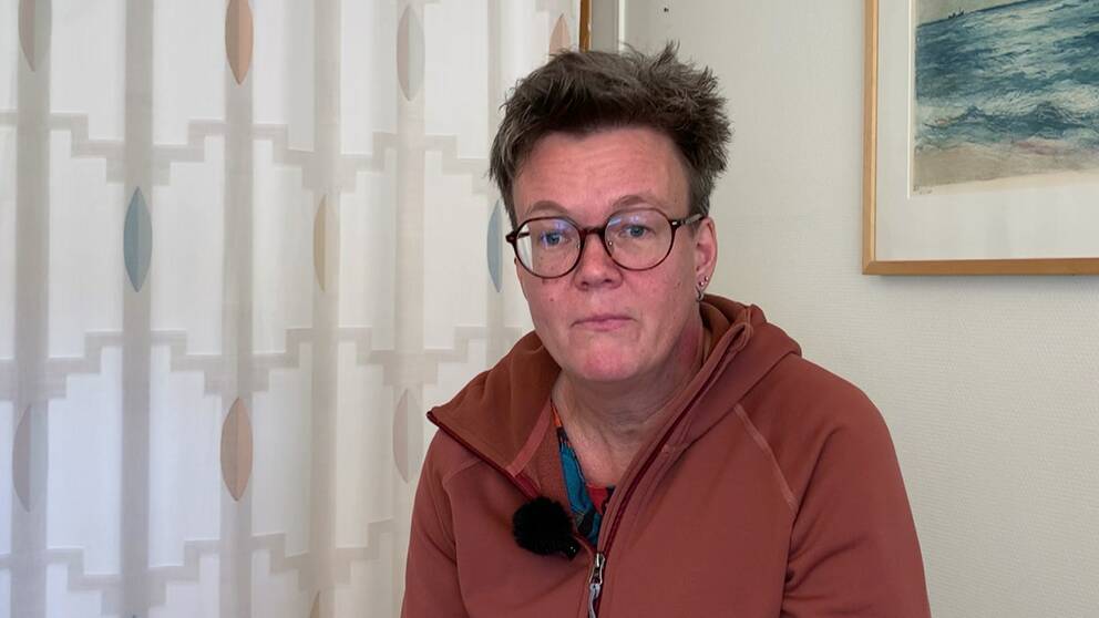 Ulrica Brandt har kortklippt mörkblondt hår. Hon har på sig en oragne hoodie och svarta glasögon. Hon sitter i ett ett vitt rum med en gardin i bakgrunden som är vit med prickar i olika färger.