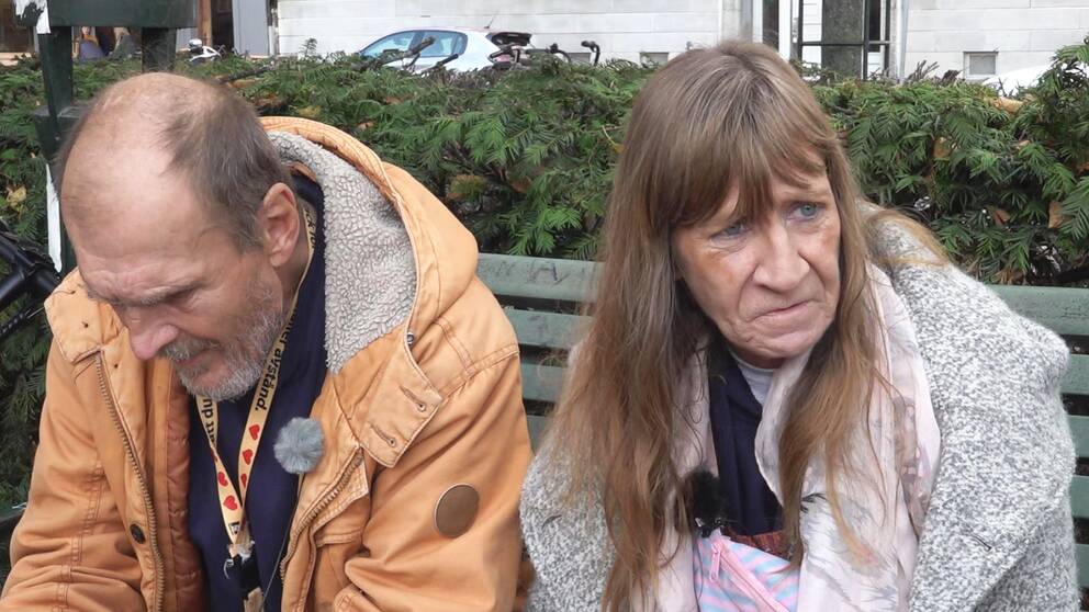 En man i orange jacka. En kvinna med lugg. De sitter på en bänk på Värnhemstorget i Malmö.
