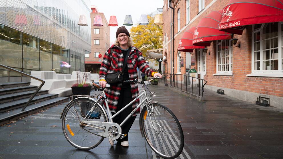 Helbild på kvinna, blont hår, brun mössa, glasögon, kappa i i rött och vitt, står och håller sin cykel på en gågata med grå stenplattor.