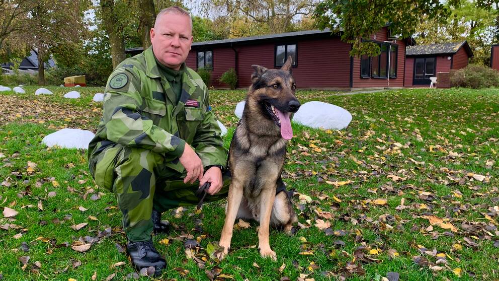 Pierre Wahlström, sitter iklädd en grön kamouflagemönstrad uniform och ser bestämd ut. Bredvid honom sitter en schäfer med tungan ute ur munnen och ser ivrig ut.