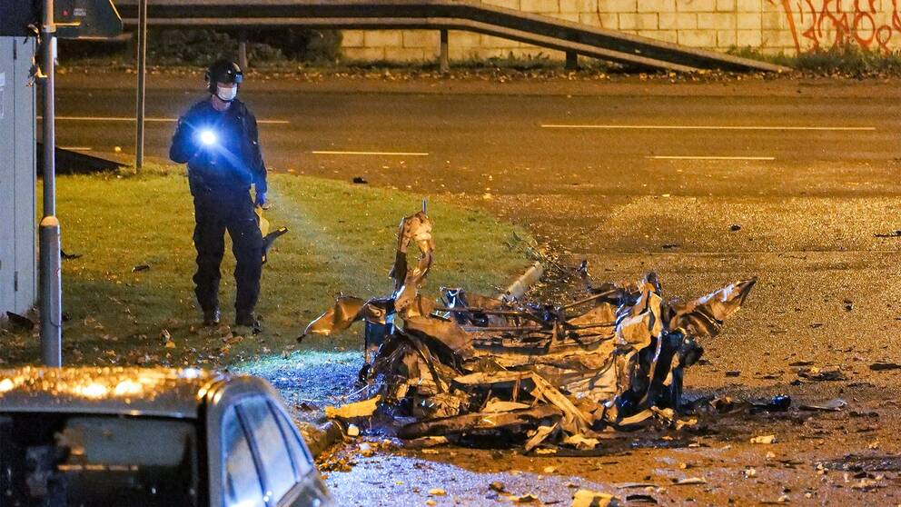 En polis står och lyser med en ficklampa på en ett förvridet skrotskelett från en bil.
