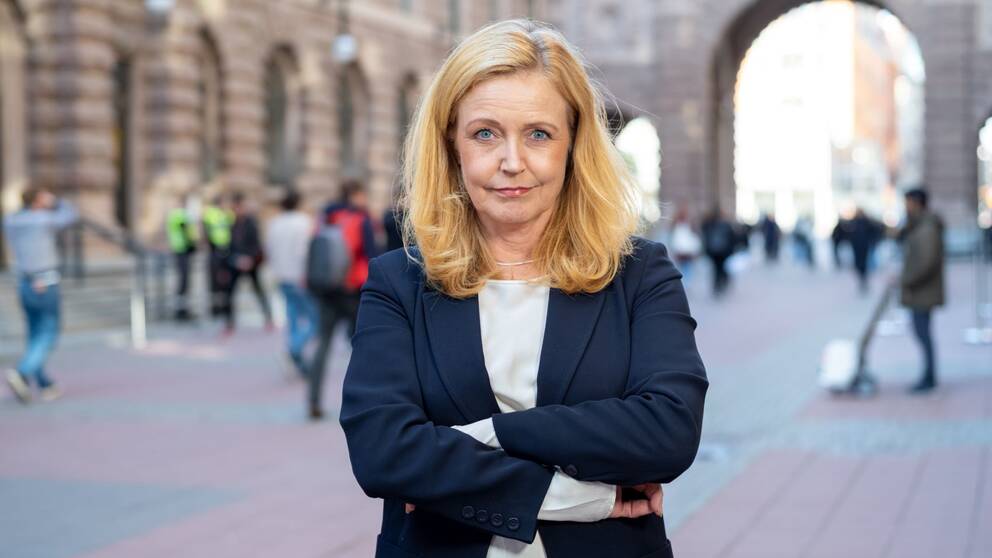 En kvinna med långt ljust, rödlätt hår i kavaj och med armarna i kors utanför riksdagshuset i Stockholm.