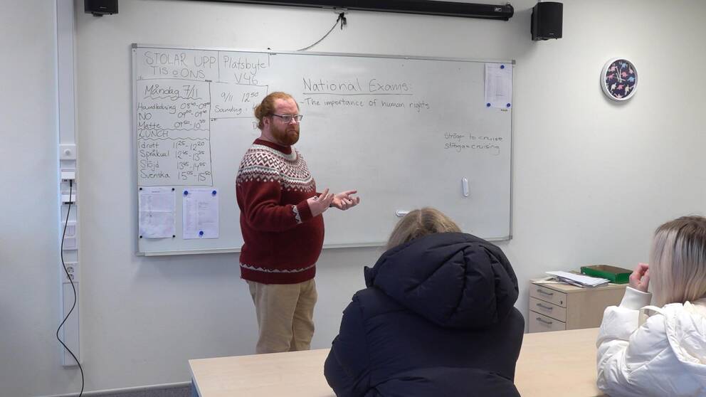man i stickad tröja, står framför en vit tavla, i ett klassrum, han har skägg och glasögon, jobbar som lärare