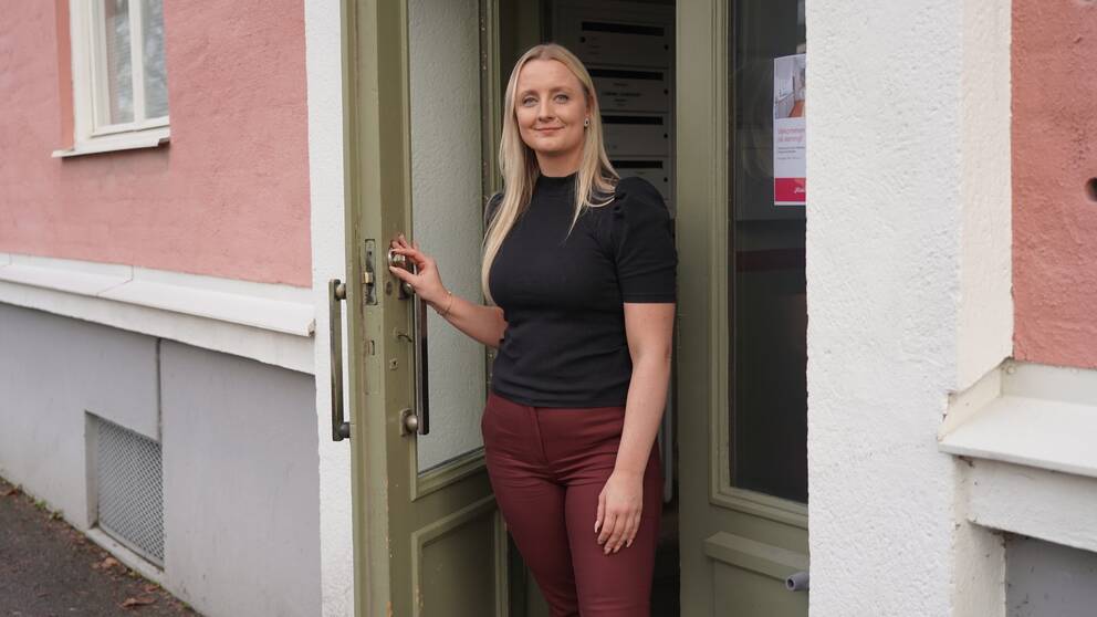 Kvinna står och håller upp en dörr, hon har blont hår, svart t-tröja och vinröda byxor. Dörren är grön och har avlånga glasrutor, huset är rosa.