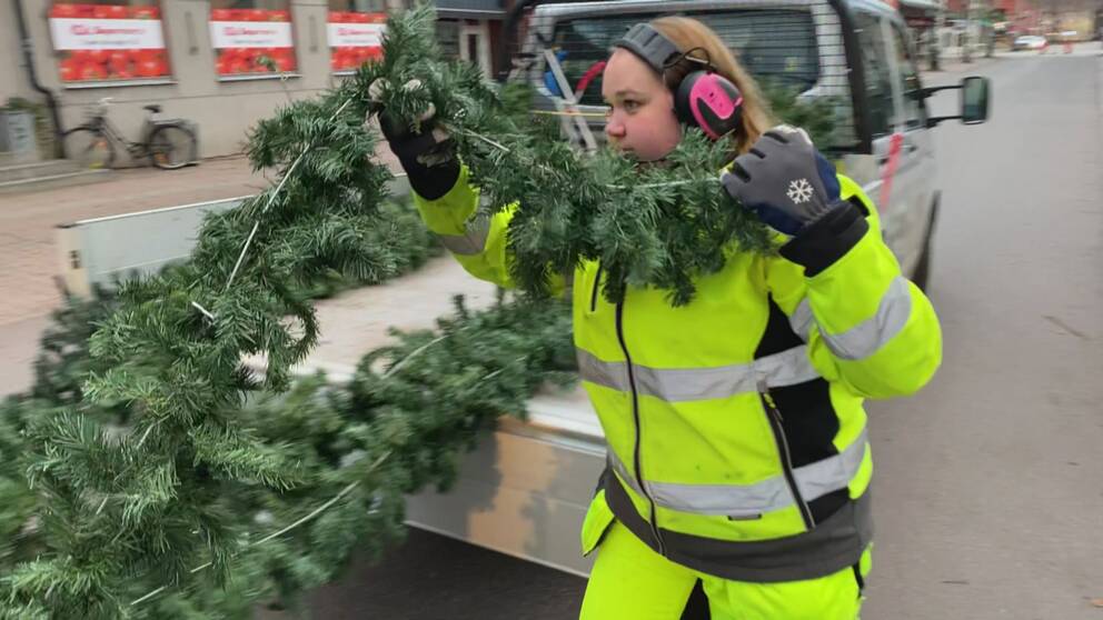 en kvinna med jobbkläder, reflexoverall och hörselkåpor och handskar, bär en granjirlang från ett flak, på en gata i Rättvik.