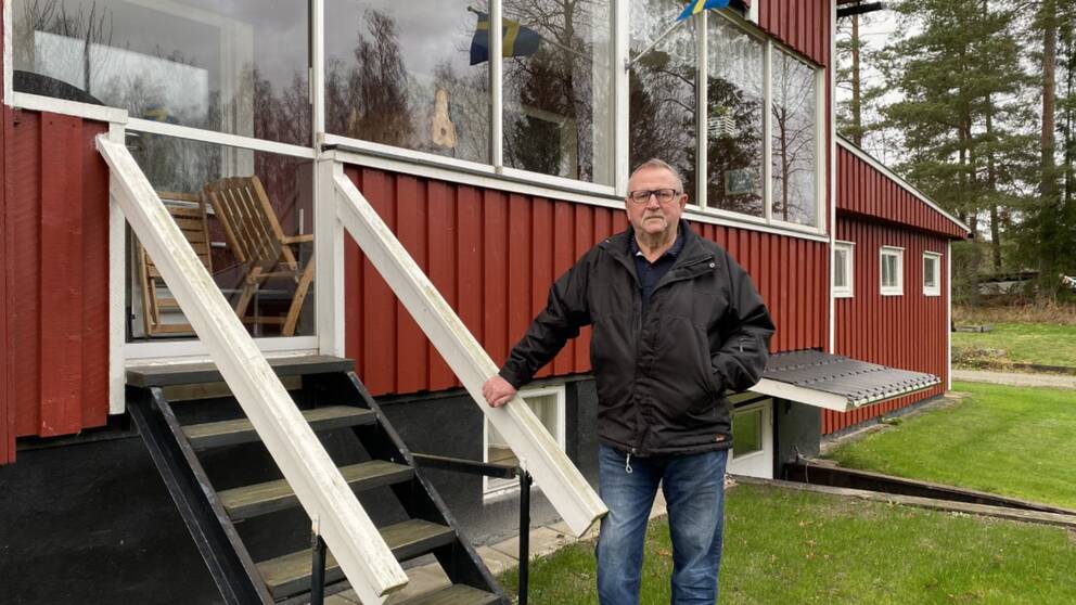 En man med glasögon står vid att trappa framför ett rött hus.