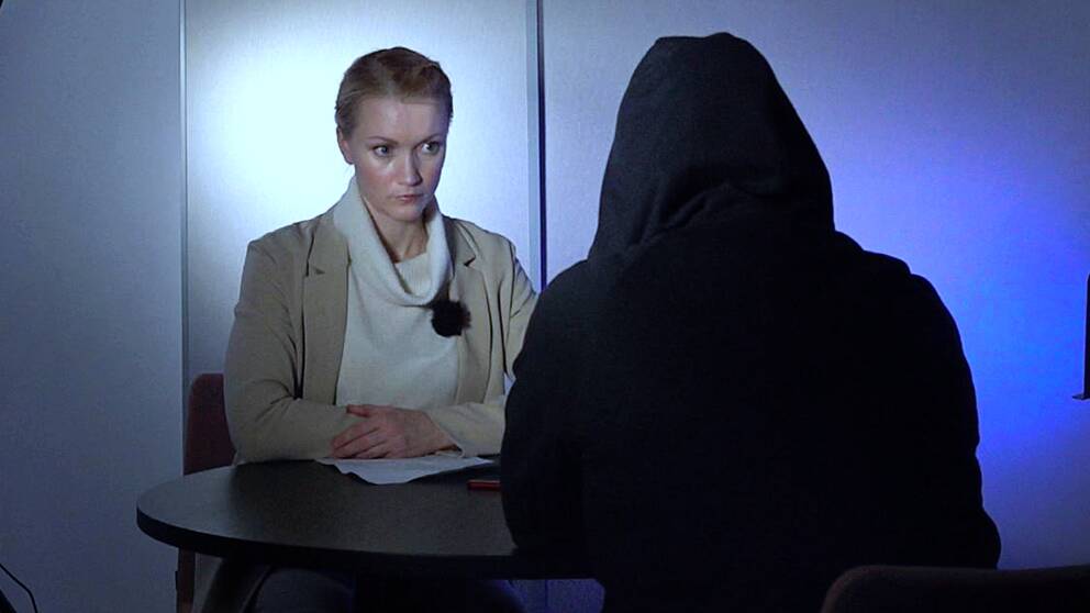 en ung kvinnlig reporter sitter vid ett bord med kvinnan som är klädd i huvtröja för att inte gå att känna igen