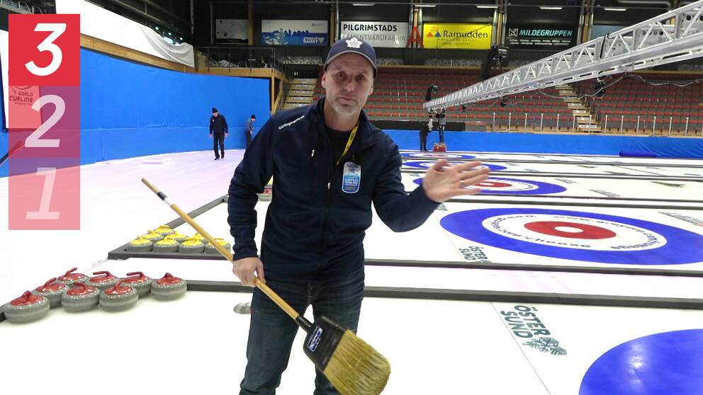 Marcus Olovsson står på en curlingbana med en sop i ena handen. I bakgrunden syns en tom läktare och personer som jobbar med förberedelser inför EM i curling.