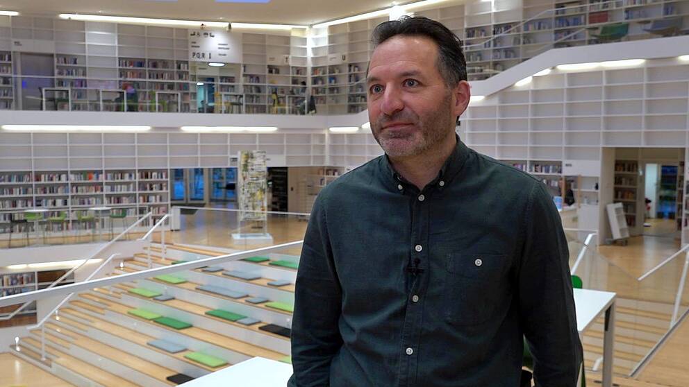 Michael Oppenheimer står i grön skjorta, högskolans bibliotek är i bakgrunden, hyllor med en massa böcker.