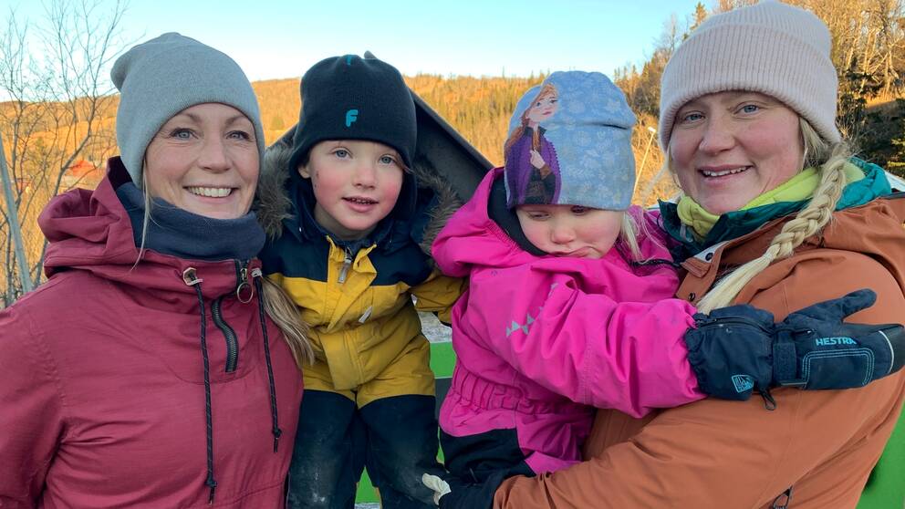 Två mammor och deras barn i Storlien, iklädda vinterkläder, tittar in i kameran och ler. I bakgrunden syns en blå himmel och berg.