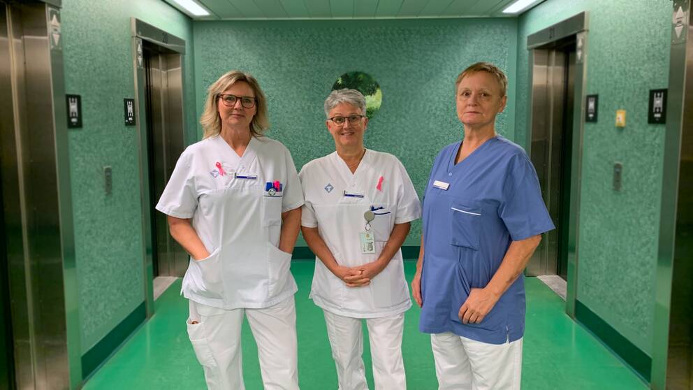 Tre sjuksköterskor uppställda i korridor på sjukhus ler mot kameran. 