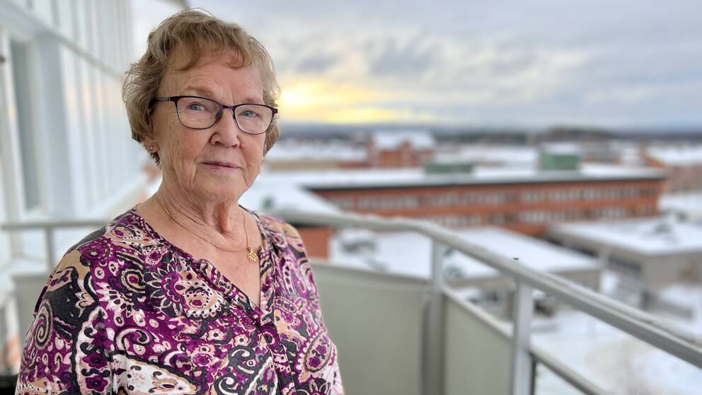 Elise Nilsson i en tunika med lila, gult och svart mönster står ute på sin balkong i Ånge. Hon har kort hår med lugg och ett par lila glasögon i tunna metallbågar. I den blurriga bakgrunden ser man vintertäckta tak på husen runtomkring.
