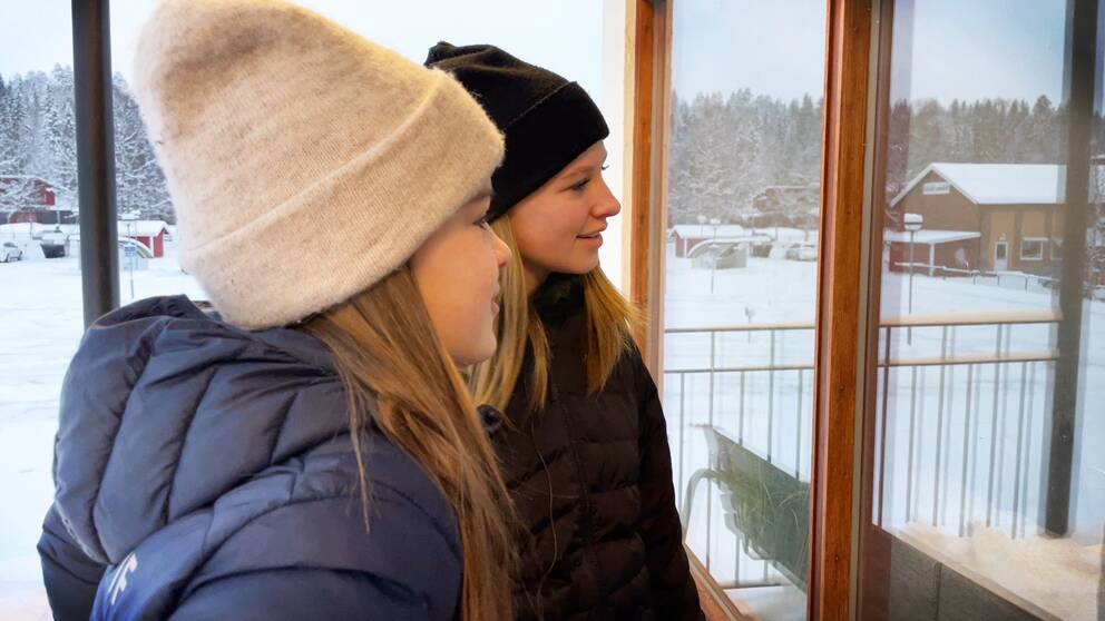 Två unga tjejer tittar in i en lokal genom ett fönster. Den ena har svart mössa, den andra har vit.
