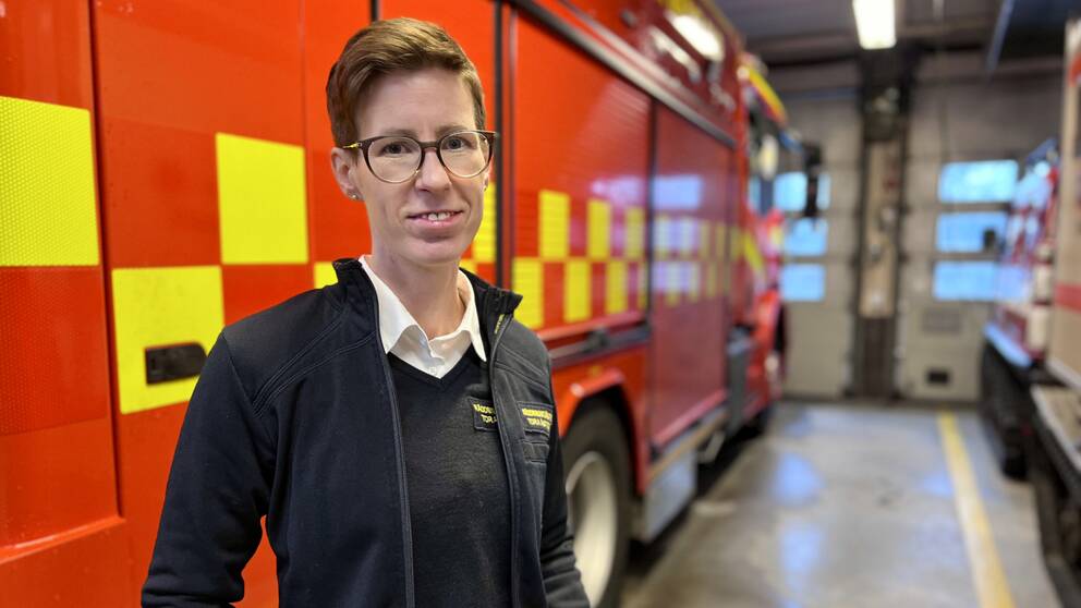 Bild på Tora Åström i räddningstjänstens arbetskläder. Hon har halvrunda glasögon, kort hår, och står framför en brandbil.