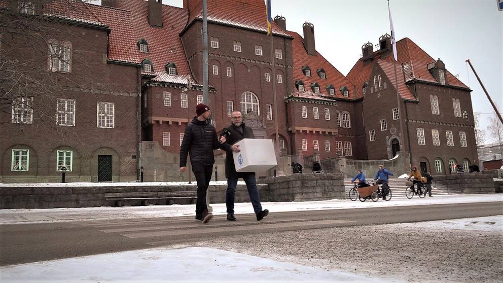 En reporter med mikrofon och en man med vit flyttkartong går över ett övergångsställe framför Östersunds rådhus.