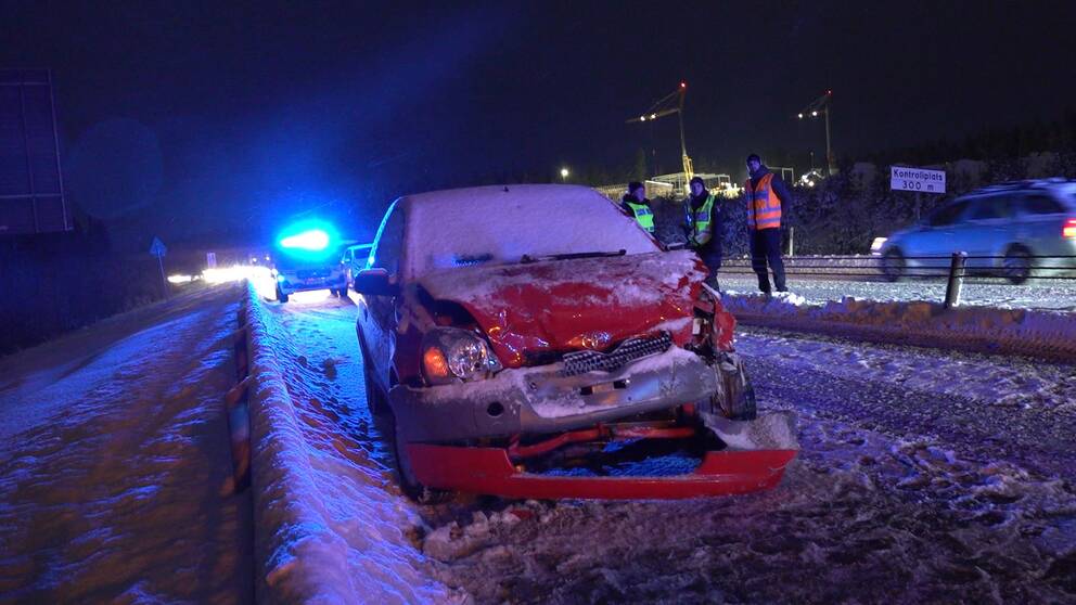 En röd, krockskadad bil står intill ett snöigt vägräcke. I bakgrunden anas poliser i varselvästar.