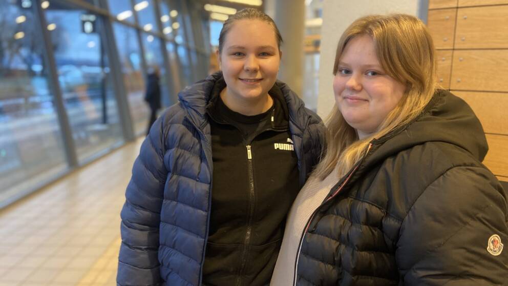 Jennelie Svensson och Linnéa Larsson står bredvid varandra i en bussterminal och tittar in i kameran.