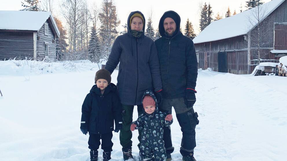 En mamma, pappa, son och dotter. I bakgrunden ser man en ladugård och ett gammalt timmerhus. Det ser kallt ut och det är mycket snö på marken.