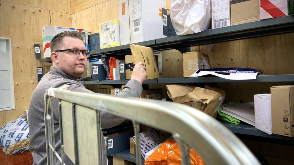 En man (Joakim Ohlsson som är postombud) står i ett postrum med en massa paket