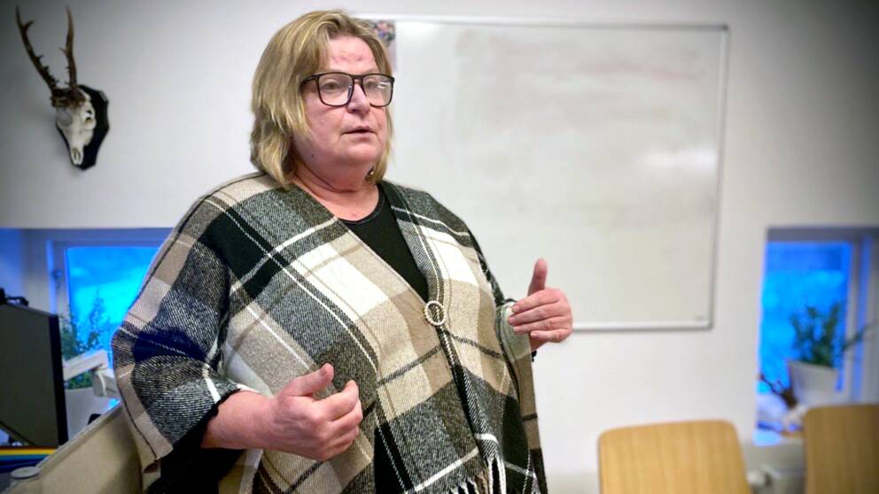 En kvinna (Margareta Jonsson tillförordnad institutionschef på Margretelund) i en poncho står i ett rum. Hon gestikulerar med händerna som att hon förklarar något