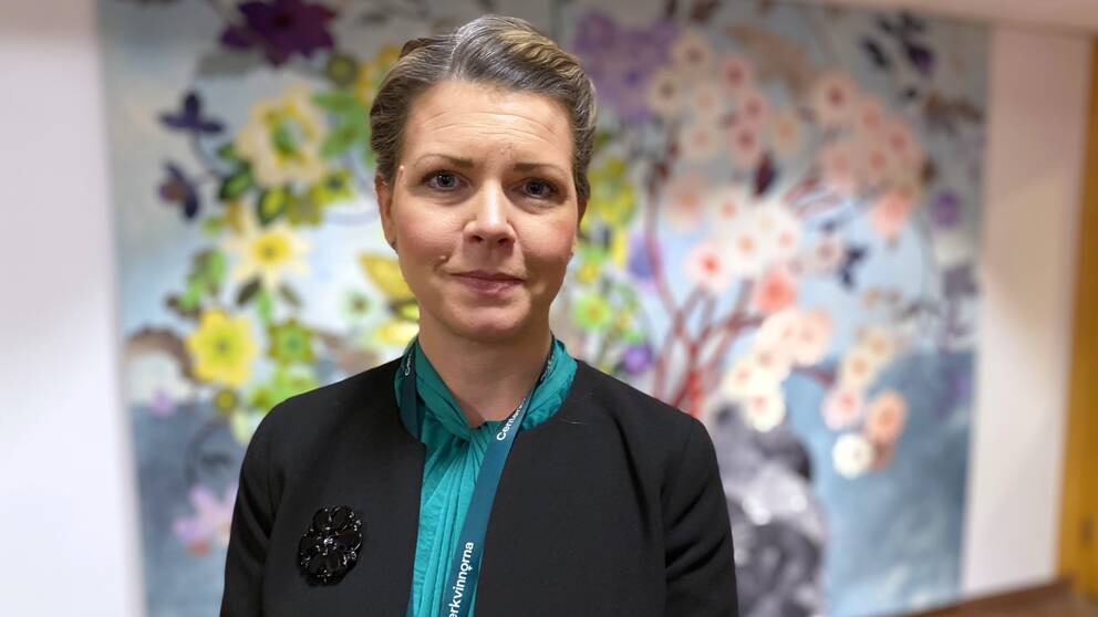 Regionrådet Sofia Jarl representerar Centerpartiet och står i entrén på regionkontoret i Falun och berättar vilken lösning hon tror på för att lösa personalkrisen inom ambulansen. Bakom henne finns en vägbonad fullt med vävda blommor.