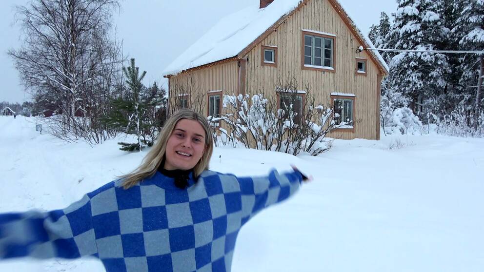 Anna Nilsson har en blårutig tröja och står med armarna utsträckta framför huset hon köpt i Gargnäs. Det syns att huset stått tomt en längre tid, med stora färgflagor i den beiga färger och de bruna husknutarna. Det är vinter och snö på marken.