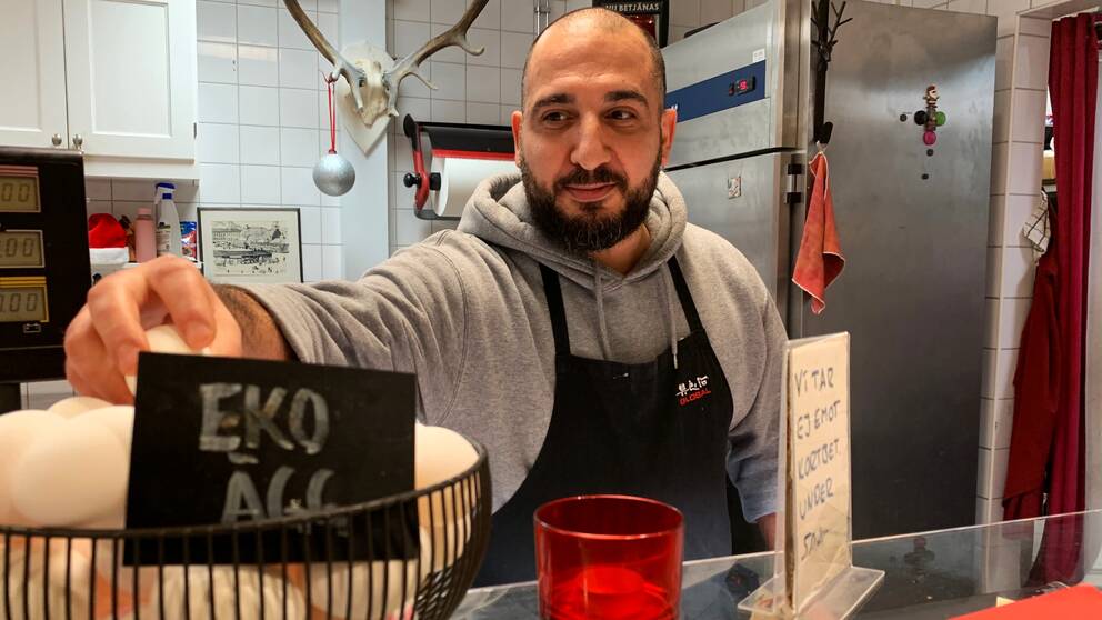 Bild på man framför restaurangkök med förklädde och gråtröja. Mannen håller i en skylt där det står eko ägg. Mannen heter  Josef Emmanuel och är en av butiksbiträdena i Saluhallen i Göteborg. Han pratar i klippet om hur inflationen påverkar