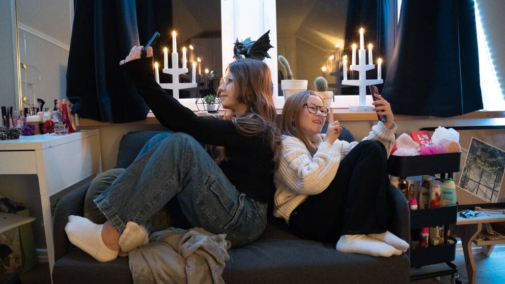 Systrarna Ann-Sofie och Linnéa Pettersson Sjørup sitter rygg mot rygg i en soffa och tittar på varsin telefon.