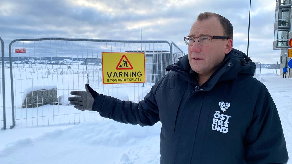 Etapp två på Storsjö Strand skulle ha varit igång nu, men det är stiltje på platsen. Bengt Gryckdal, mark- och exploateringschef i Östersund berättar varför det står stilla. På bilden ser man Bengt Gryckdal, en man med kort mörkt hår och glasögon. Han står vid ett stängsel där det hänger en gul skylt som det står ”Varning byggarbetsplats” på.