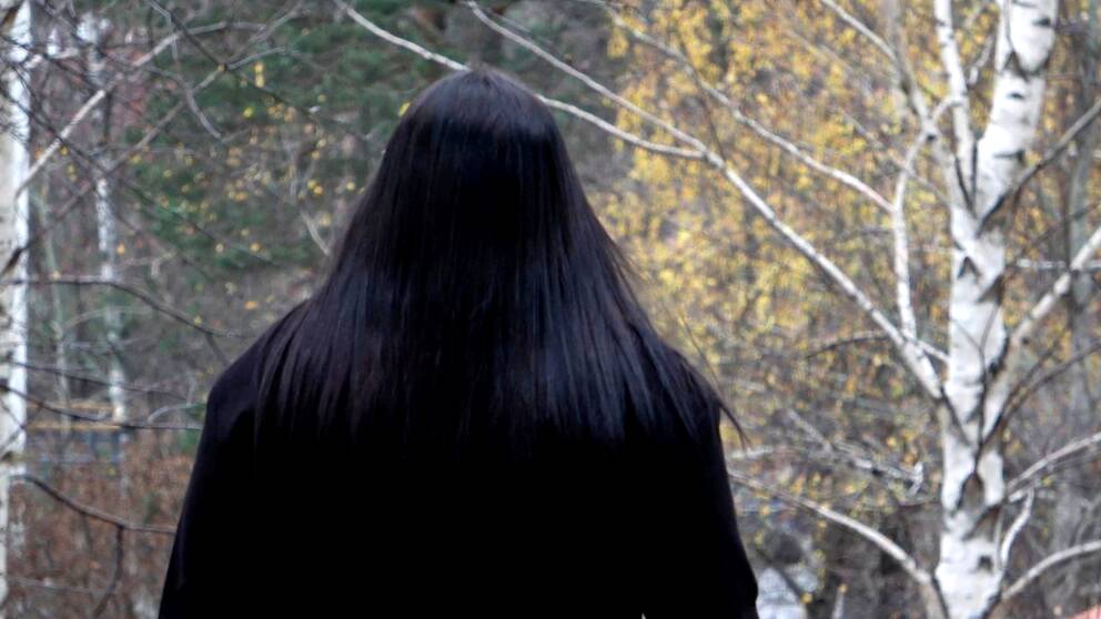 En kvinna står med ryggen mot kameran. Hon har mörkt hår och bär en mörk kappa.