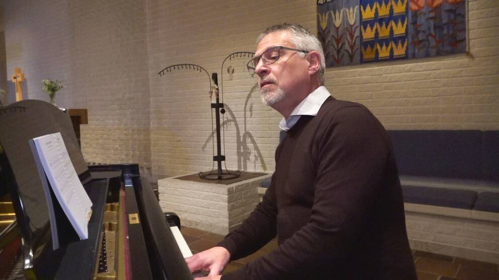 Präst sitter vid ett blanksvart piano i en kyrka och spelar samtidigt som han sjunger. 