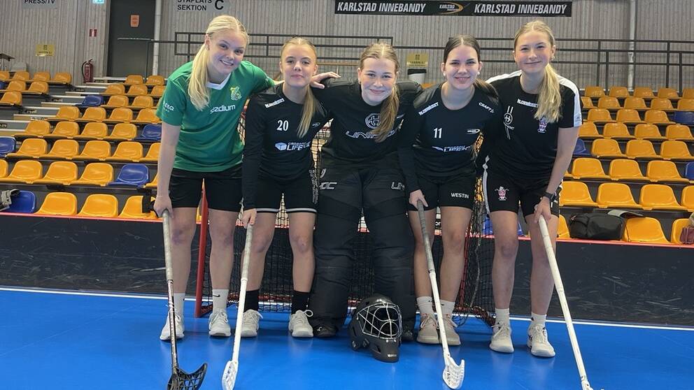 Fem unga kvinnliga innebandyspelare står på en innebandyplan med klubborna, håller om varandra och ler. Bakom dem är en läktare med stolar i gult och blått.