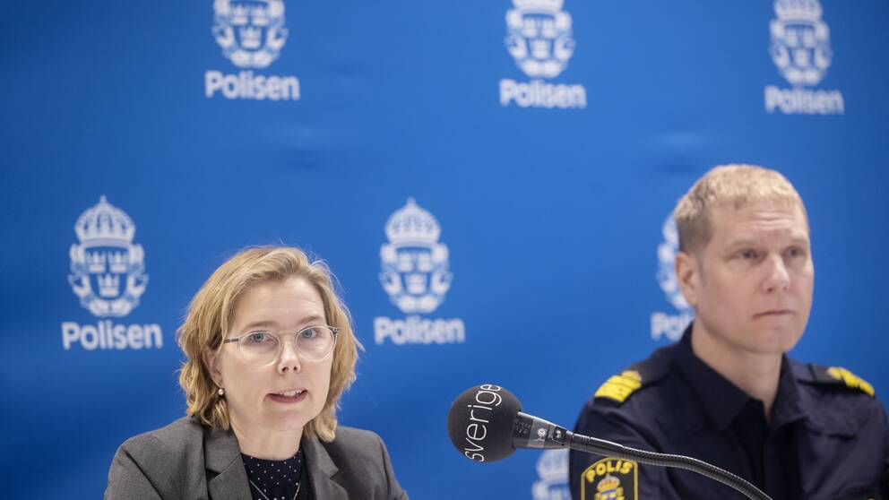 Karin Everitt har axellångt ljust hår och glasögon. Hon är iklädd svart tröja med vita prickar, grå kavaj och tittar in i kameran. Till höger om henne sitter lokalpolisområdeschefen Josef Wiklund i polisuniform. Bakom dem är en stor blå vepa med polisens logotyp i vitt.