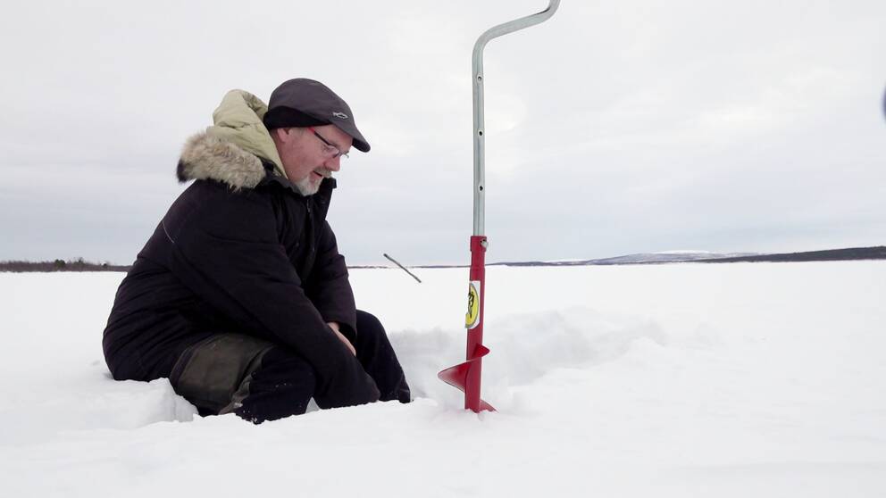 Tomas Stålnacke sitter och pimplar på isen. Framför honom står borren lite lätt fastskruvad i isen. Det är ljust och lite mulet väder, i horisonten syns en strimma land.