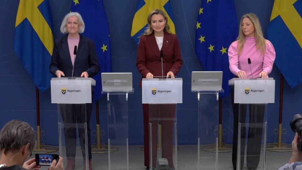 Tre kvinnor står uppradade bakom talarstolar där det står Regeringen. Bakom dem syns Sveriges och EU:s flaggor.