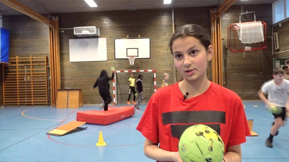 12 årig flicka i röd t-shirt som håller i en grön handboll. Bakom ser man klasskompisar som spelar handboll.