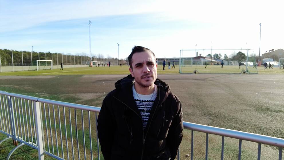 Fotbollsdomaren Sami Dalali står framför en fotbollsplan där spelare håller på med en träning. 