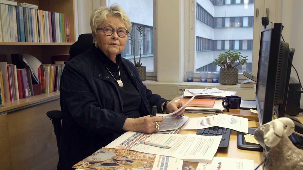 Eva Eriksson som är förbundsordförande i SPF seniorerna sitter vid sitt skrivbord. Hon har kort ljust hår, glasögon och är klädd i mörka kläder. Bakom henne finns en hylla full med böcker i olika färger och skrivbordet är fullt med olika dokument.